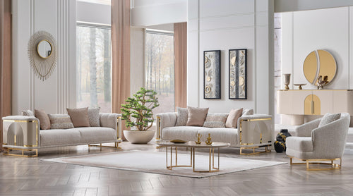 A luxury modern living room set featuring a light beige sofa, matching armchair, and a small round coffee table with gold metal legs, arranged in a bright, elegant room with beige walls, wood floors, and decorative accents.